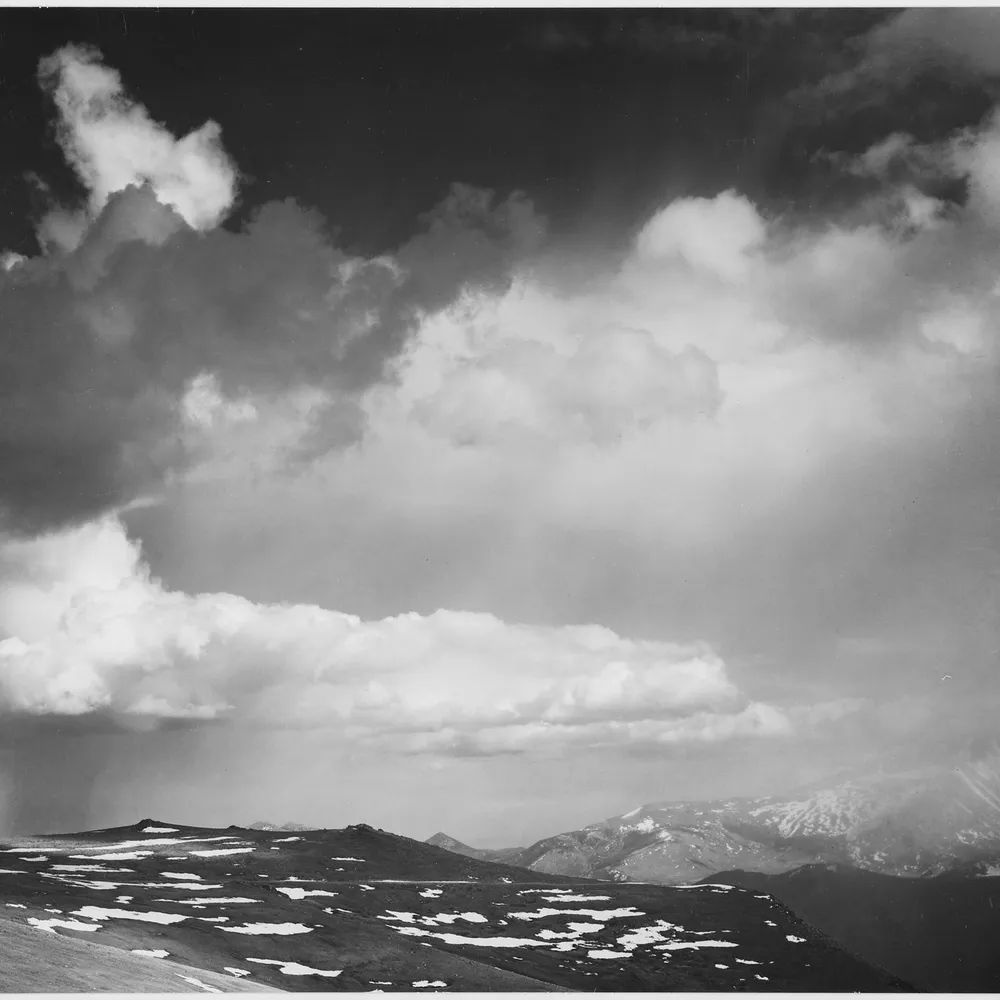 Black-and-white photograph of jagged mountain peaks beneath a near-black sky filled with bright, billowing clouds, showing the dramatic tonal separation produced by a red filter.